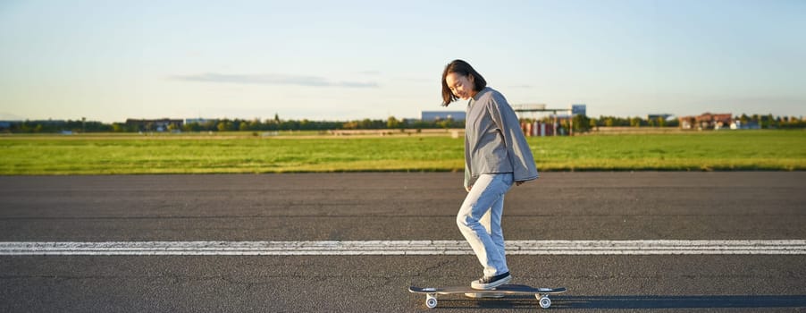 Beautiful asian skater girl riding her longboard on sunny empty road. Young woman enjoying her skate ride smiling and laughing.