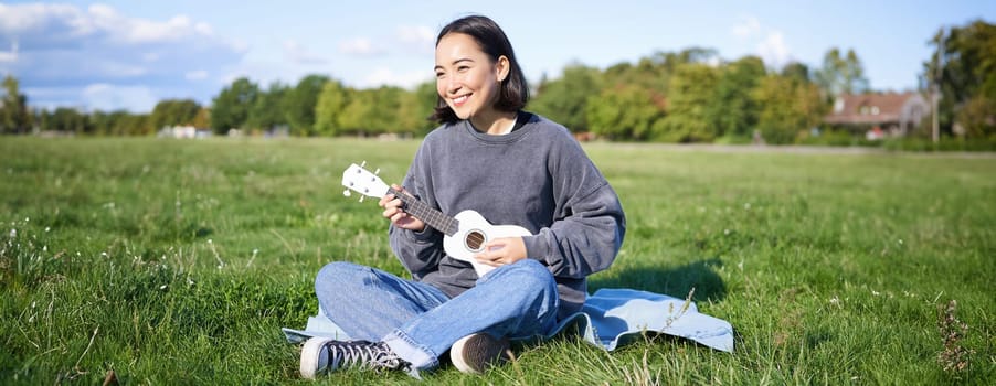 Beautiful asian girl sitting in park, playing ukulele and singing, relaxing outdoors on sunny spring day.