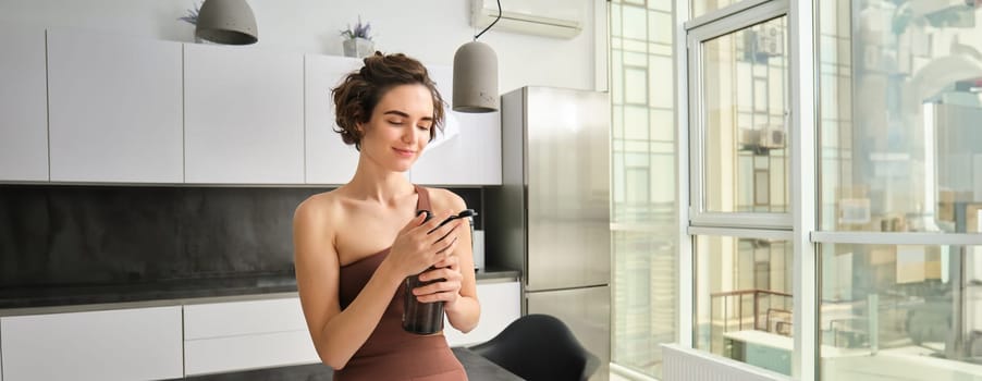 Portrait of smiling fitness girl, wearing sportswear for jogging, workout training, standing in kitchen and drinking water from sports bottle, looking fit and happy.