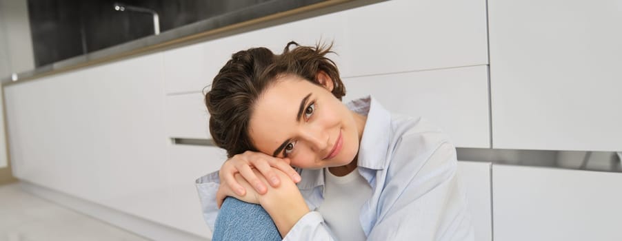 Close up portrait of beautiful, tender young woman smiling, looking at camera, sitting in cozy pose on kitchen floor, daydreaming.