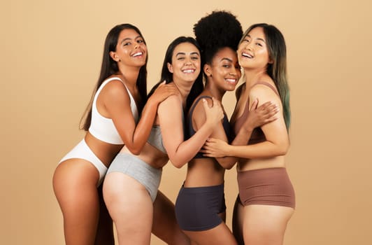 Four radiant women from diverse backgrounds pose together, laughing and embracing, flaunt their natural beauty in comfortable underwear against beige background