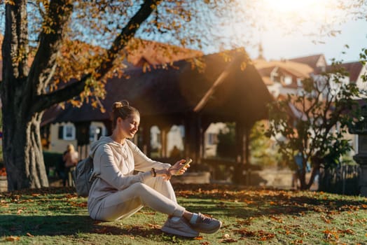 Young fashionable teenage girl with smartphone in park in autumn sitting at smiling. Trendy young woman in fall in park texting. Retouched, vibrant colors. Beautiful blonde teenage girl wearing casual modern autumn outfit sitting in park in autumn. Retouched, vibrant colors, brownish tones.