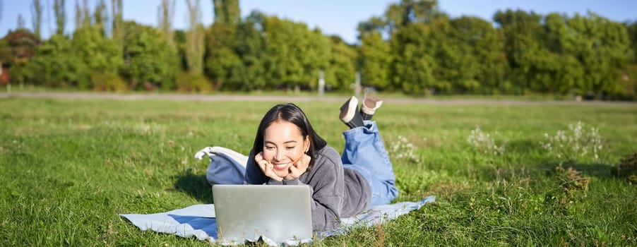 Smiling asian girl lying in park on grass, watching movie or video on laptop, looking at screen with interest.