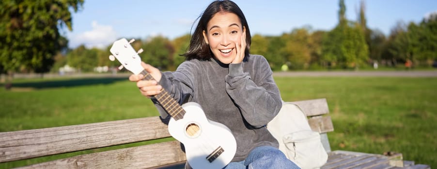 Portrait of young girl musician, sitting in park with ukulele guitar, looking surprised at camera, saying wow.