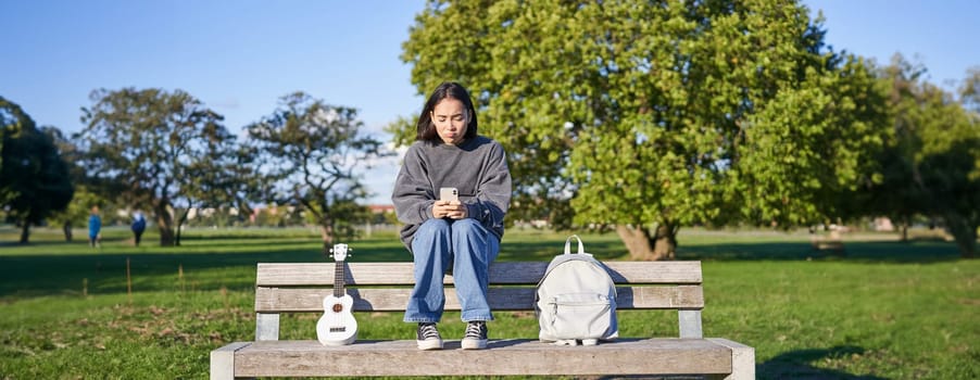 Beautiful brunette girl on bench in park, sitting with ukulele and backpack, holding smartphone, using mobile app.