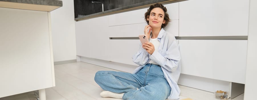 Portrait of girl with mobile phone, sits on floor at home, orders takeaway in app. Young woman with smartphone does online shopping from home, browsing websites.