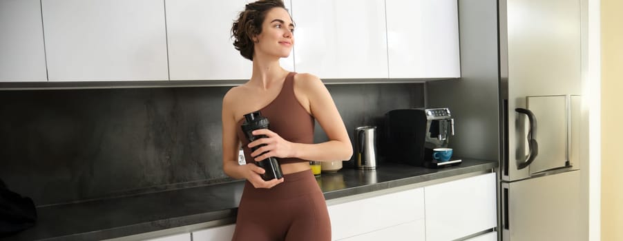 Portrait of smiling fitness girl, wearing sportswear for jogging, workout training, standing in kitchen and drinking water from sports bottle, looking fit and happy.