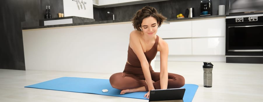 Portrait of smiling young woman, fitness instructor on rubber yoga mat, joins online training class from tablet, listens video in wireless headphones, workout at home on floor.