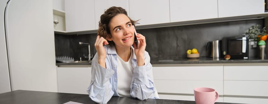 Smiling young woman talks on mobile phone, calls someone from home, sits in kitchen and has conversatiopn