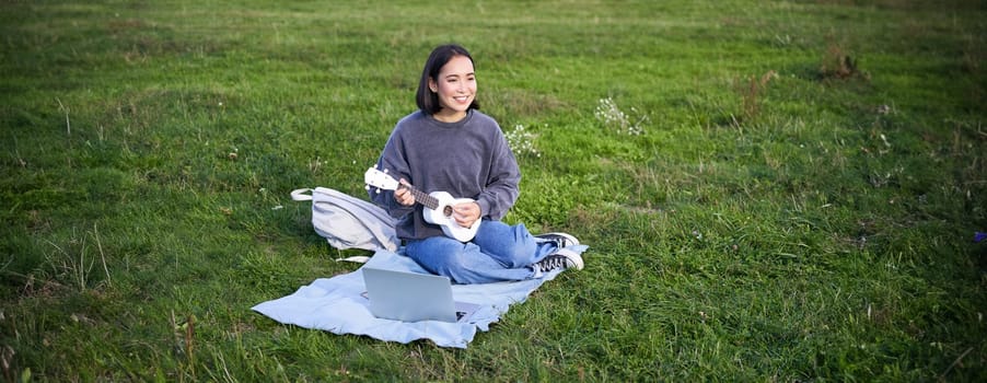 Cute korean girl, musician sits in park, plays ukulele and sings, looks up chords and tutorials on laptop.
