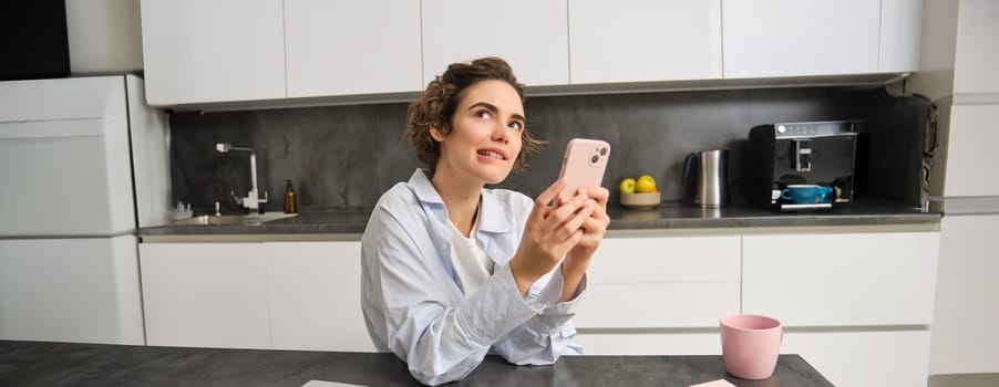 Portrait of woman thinking while holding smartphone, deciding what to order on mobile phone app.