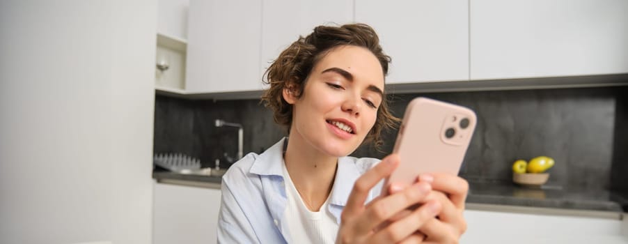 Portrait of modern brunette woman holding mobile phone and smiling. Girl sits in kitchen at home with smartphone, using app to order delivery, does online shopping.