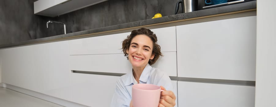 Portrait of happy cute woman gives you cup of coffee, offers to drink tea, sits on floor in kitchen and smiles.