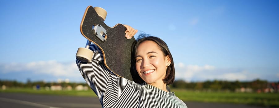 Happy and free asian girl holding cruiser board on shoulders and walking towards camera on empty road, skating on longboard and enjoying sunny weather.