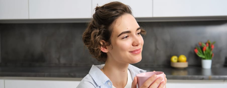 Happy brunette woman drinking coffee at home. Girl with pink cup, sits in her kitchen and enjoys relaxing morning weekend.