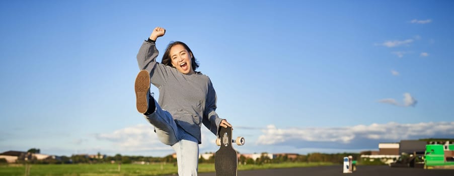 Excited asian girl dancing, standing with skateboard, skating on longboard and shouting carefree.