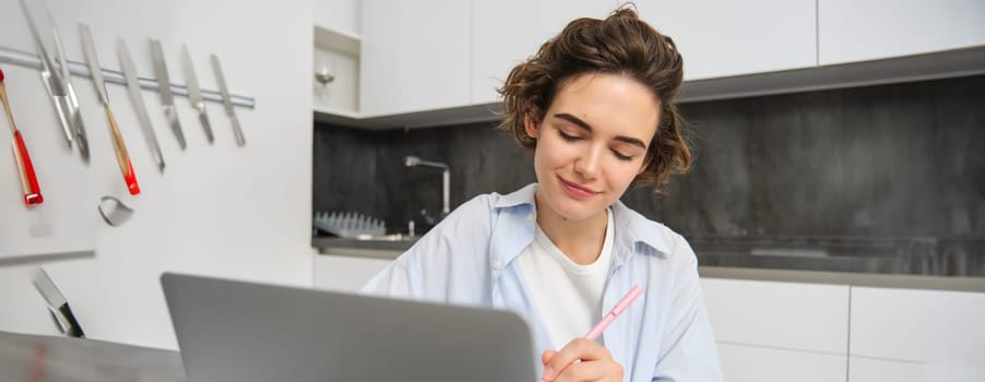 Image of young woman, entrepreneur works from home. Girl studies remote, writes down information, makes notes, uses laptop, does homework online.