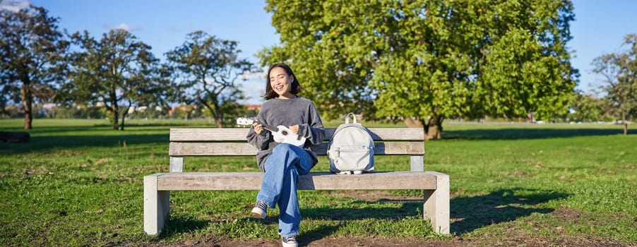 Carefree girl sits on bench in park with ukulele, plays and sings outdoors on sunny happy day.