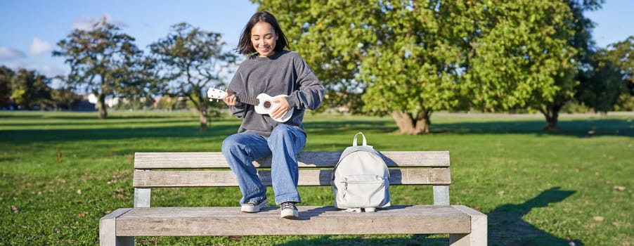 Portrait of young hipster girl sits on bench and plays ukulele, sings along, relaxing in park with her musical instrument.