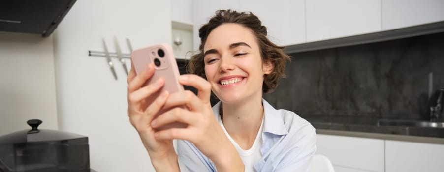 Portrait of brunette woman chats on mobile phone, uses smartphone app from home, orders delivery while sits in kitchen.