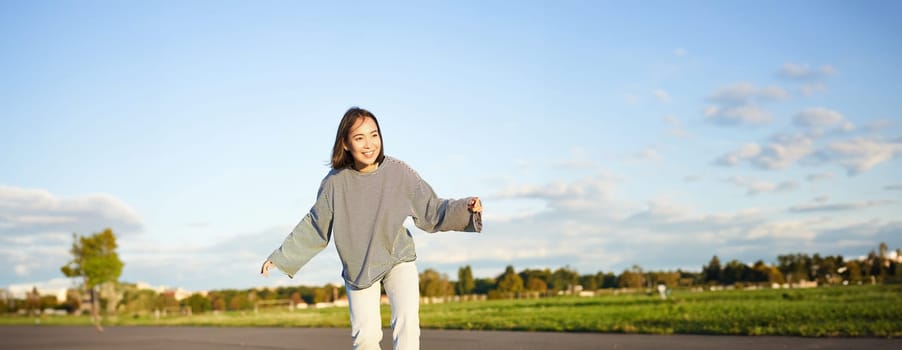 Cute asian girl riding skateboard, skating on road and smiling. Skater on cruiser longboard enjoying outdoors on sunny day.