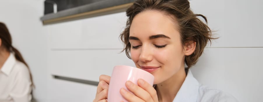Beauty and women concept. Relaxed young woman enjoys her cup of tea. Girl sits on floor in kitchen with pink mug, drinks coffee and smiles from delight.