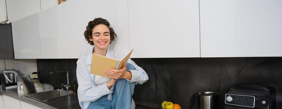 Lifestyle and people concept. Young woman enjoying weekend at home, reading notes in her journal, sits on kitchen counter with notebook and smiling.