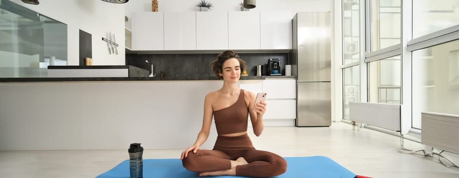 Sport app and wellbeing. Young woman, athlete sitting on floor at home with smartphone, doing yoga, using workout app on mobile phone, doing exercises at home. Copy space