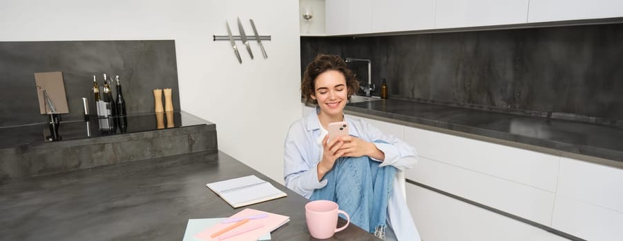 Young woman having break from work, sitting in kitchen and using smartphone, girl studies at home, sends a message on mobile phone.