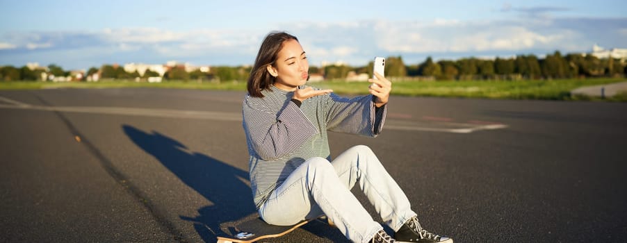 Happy asian girl sits on skateboard, takes selfie with longboard, makes cute faces, sunny day outdoors.