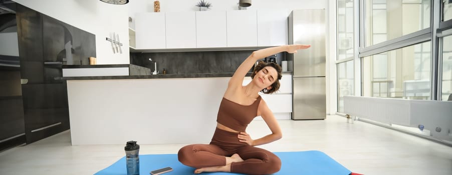 Fitness girl doing exercises at home. Young woman athlete sitting on yoga mat in bright room, stretching her arms, doing pilates exercises. Copy space