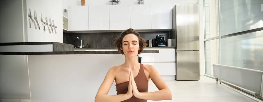 Close up portrait of young woman at home, sitting and meditating, practice yoga, making lotus pose, exercise indoors. Sport and women wellbeing concept