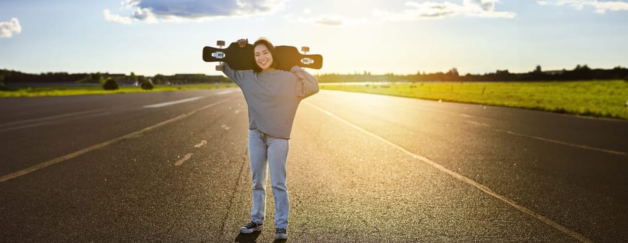 Young asian woman standing with longboard on sunny road, skating in skate park on her cruiser. Copy space