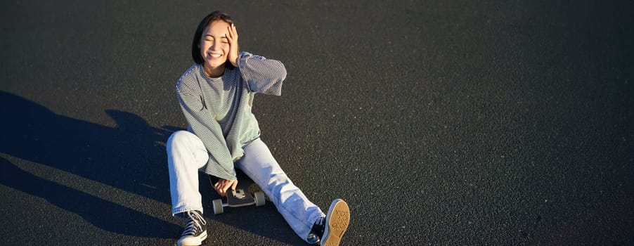 Beautiful korean teen girl sits on skateboard and enjoys sunny day. Smiling young asian woman skating on longboard.