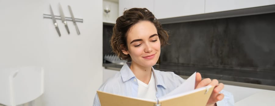 Young woman sitting in kitchen, reading her notes in notebook. Smiling girl at home, doing homework, studying.