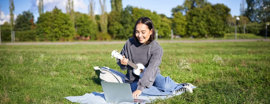 Happy asian girl plays ukulele outdoors, teaches music online with her laptop, sitting in park, playing instrument.