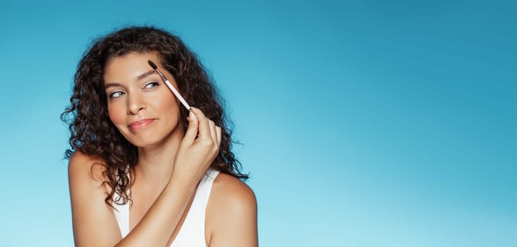 Positive millennial european curly lady with brush combing eyebrows, makes base makeup, isolated on blue studio background, panorama. Beauty care and daily routines, fashion
