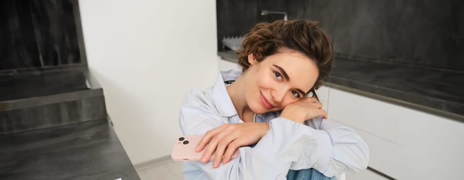 Feminine smiling girl sitting on kitchen chair with smartphone, daydreaming, looking happy. Mobile connection and people concept