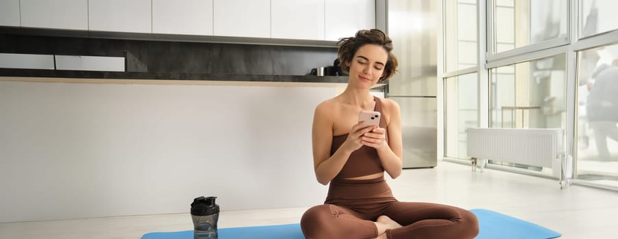 Yoga and sport at home concept. Young woman sitting in bright rome with mobile phone, doing workout on rubber mat, searching for exercises online on smartphone app, using sports equipment.