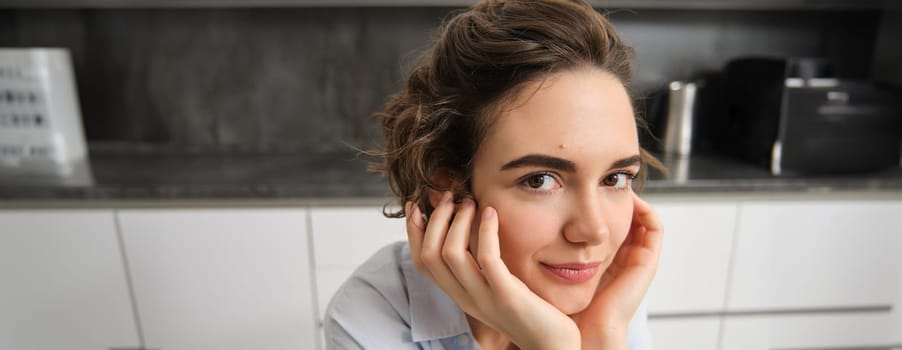 Close up portrait of young woman, 25 years old, sitting in her kitchen alone, express candid happiness, smiles, leans head on hands.