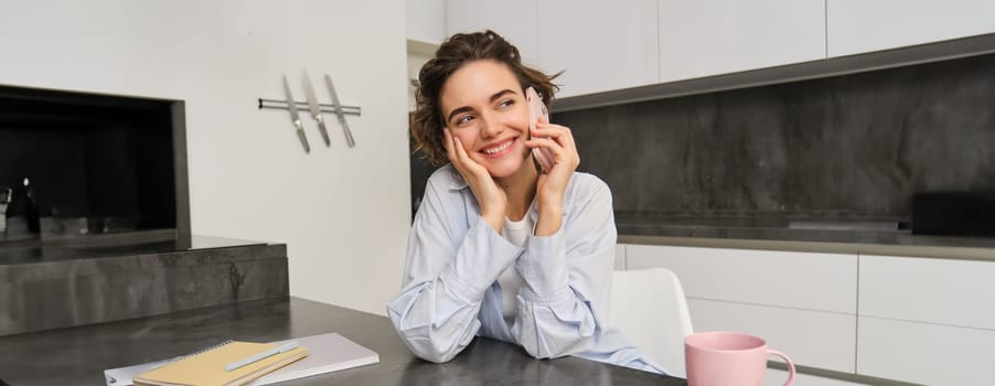 Portrait of young woman, 25 years old, holding smartphone, talking on mobile phone, sitting in kitchen at home.