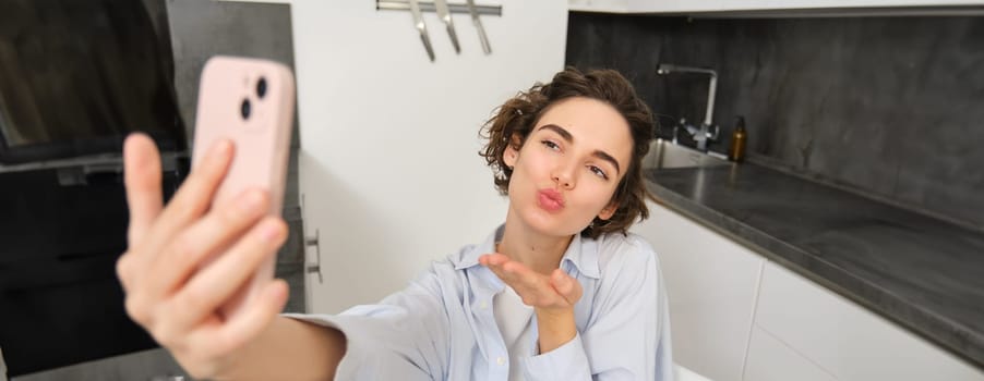 Portrait of cute brunette woman takes selfie, sends kisses at smartphone camera, records video on mobile phone app.