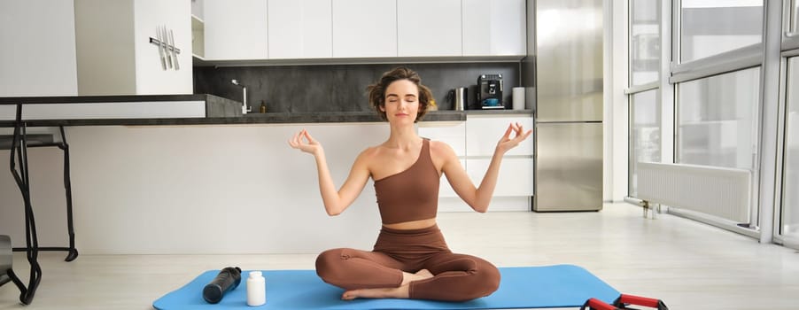 Portrait of young fitness woman, female athlete practice yoga from home, sitting in lotus pose with relaxed face, using rubber mat in kitchen for workout. Sport and wellbeing concept