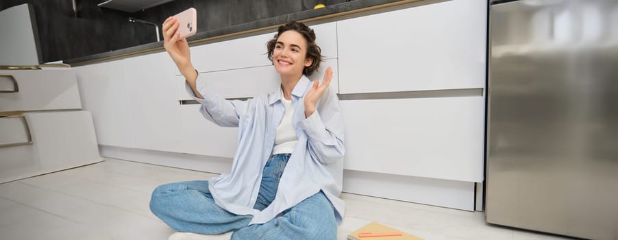 Portrait of young woman video chats, records vlog while sits on kitchen floor. Cute girl takes selfie on smartphone.