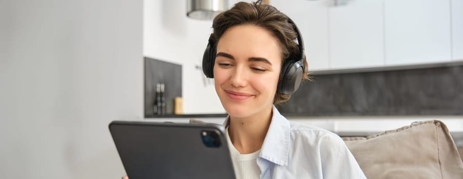 Portrait of freelancer girl sits with headphones, smiles and looks at digital tablet, works from home. Copy space