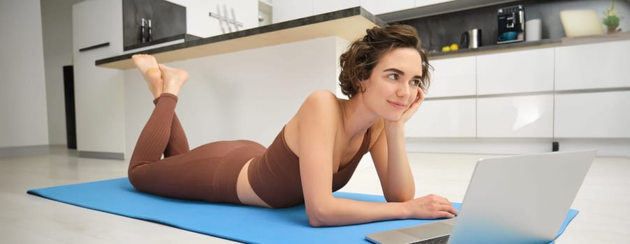 Portrait of woman athlete, girl workout at home, watches fitness videos on laptop, lying on rubber mat and doing indoor yoga session. Sport and wellbeing concept