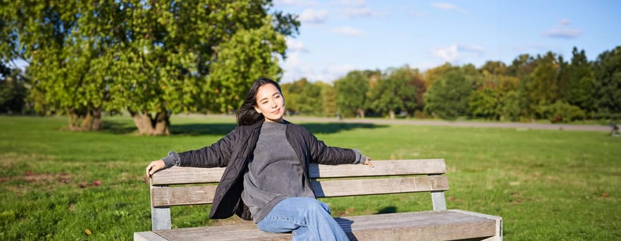 Portrait of young woman in outdoor clothes, sitting on bench relaxed, smiling and enjoying view on green park.