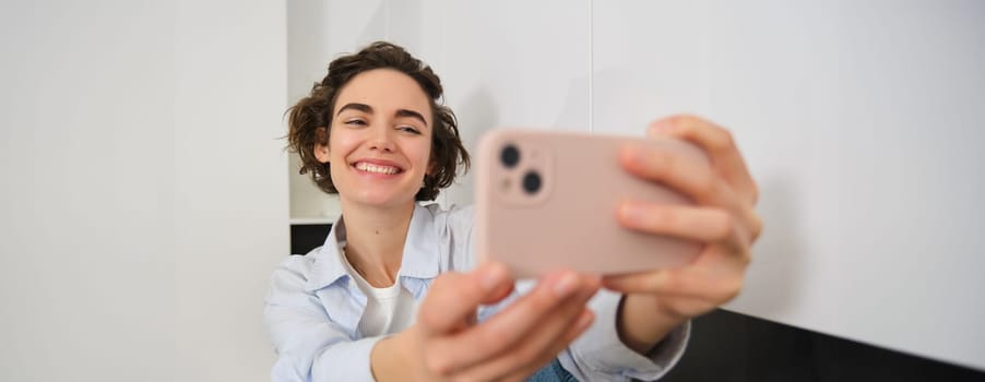 Portrait of beautiful brunette girl, takes selfie on smartphone in her kitchen, posing for photo on mobile phone.