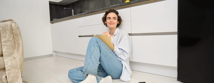 Portrait of cute brunette woman sits on floor, draws in her planner, writes diary on floor in kitchen and smiles. Leisure, lifestyle and people concept