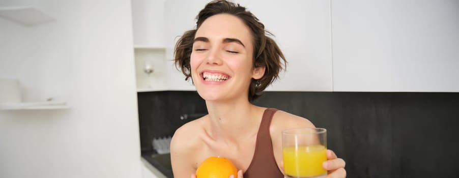 Healthy lifestyle and sport. Beautiful smiling woman, drinking fresh orange juice and holding fruit in her hand, enjoying her vitamin drink after workout at home, standing in kitchen.